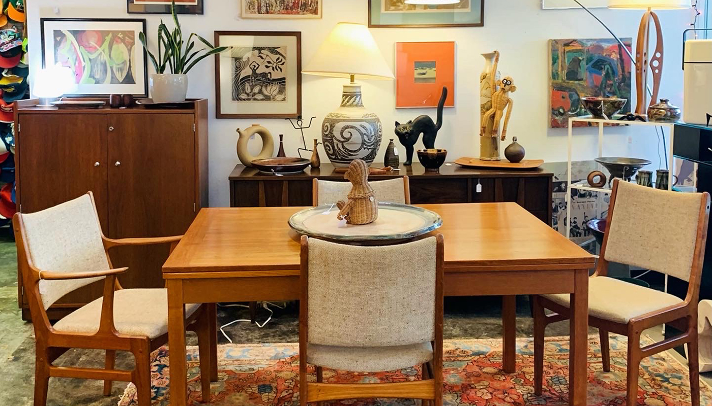 Mid-century modern wood dining table and chairs in a store. The wall in the background features many pieces of art.