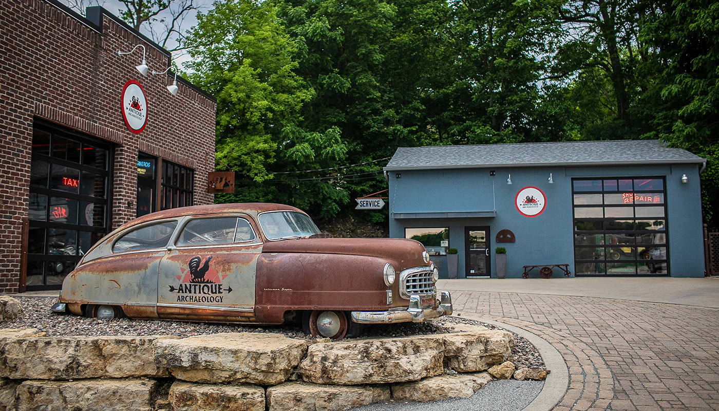 A rusted vintage car parked outside of a shop, surrounded by a brick driveway. A blue building is on the right.. 