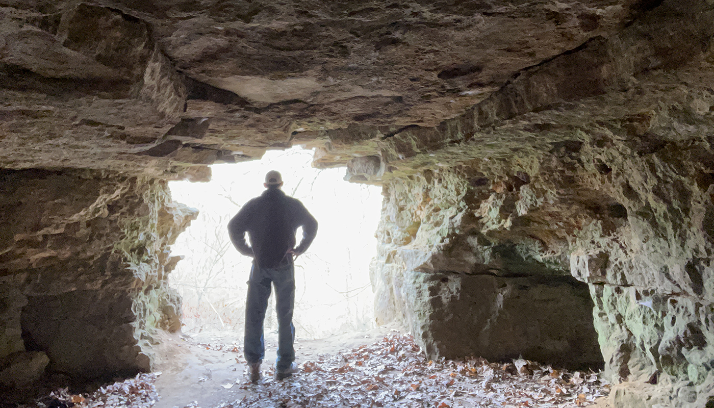 A man stands in the mouth of a cave, looking out into the sunlit woods.
