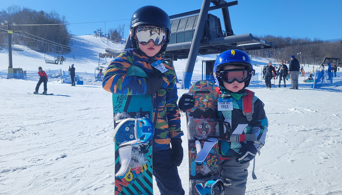 Two kids in snowsuits, helmets and goggles stand with their snowboards at the bottom of a ski slope. Various people are snowboarding behind them