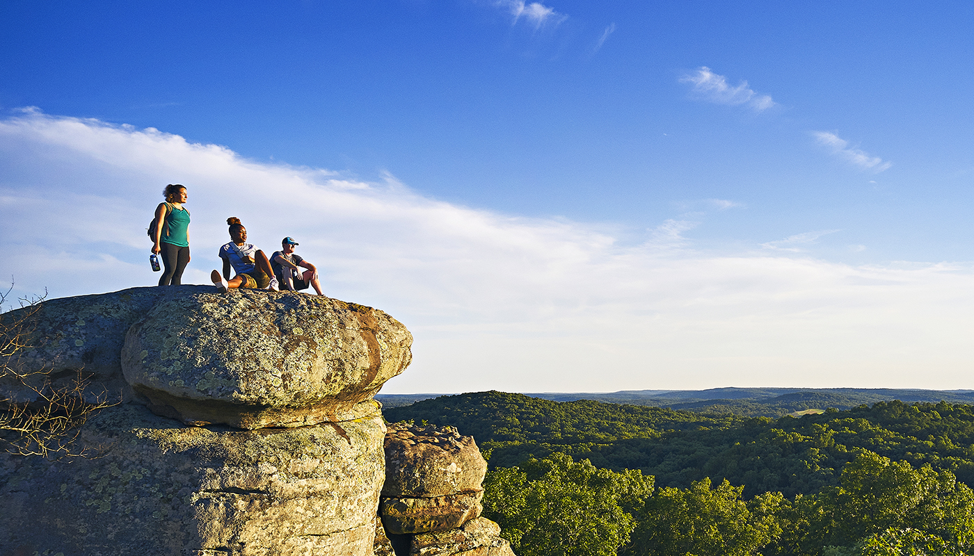 Three hikers rest on a rock formation with a view of the forest below. 