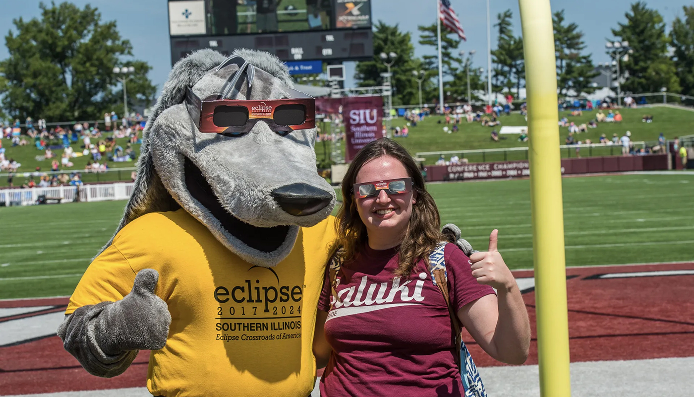 A woman poses with a thumbs up next to a dog mascot on a football field.