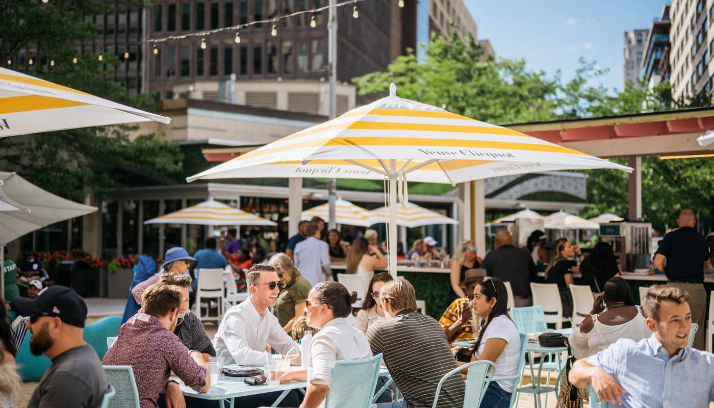 Patrons enjoy dining in a sunny outdoor eating area. Some tables have umbrellas.