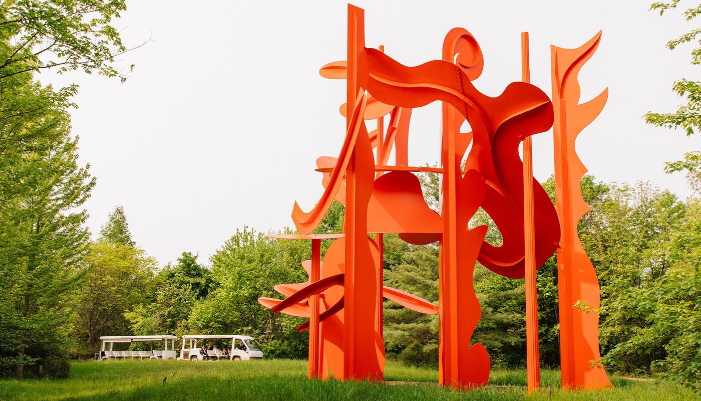 An abstract orange sculpture in front of a background of green trees. 