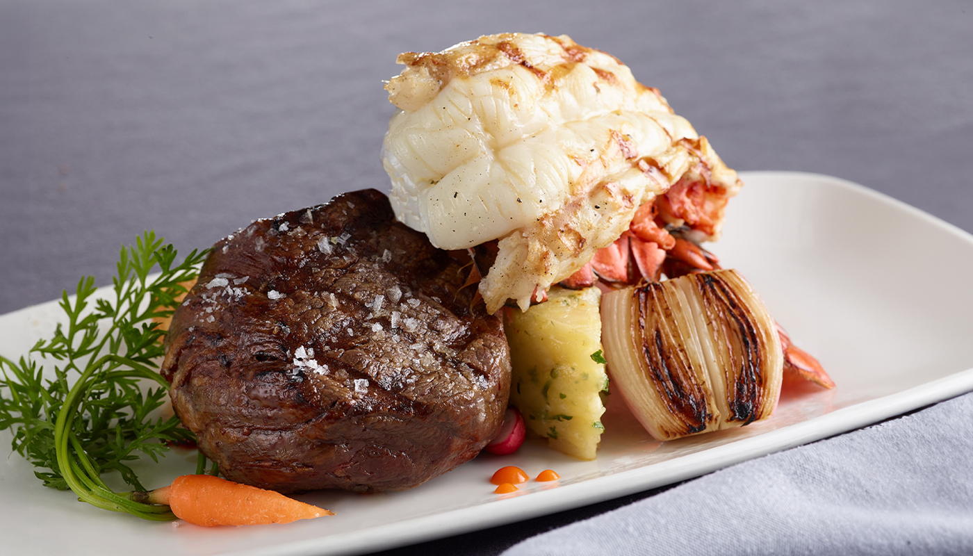 A plate of filet mignon and lobster tail, garnished with parsley and baby carrots
