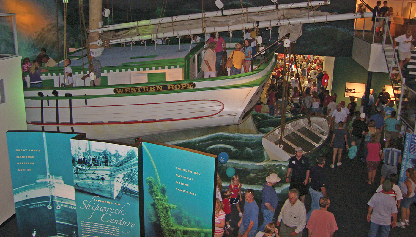Visitors gather around the exhibits of the Great Lakes Maritime Heritage Center. One exhibit is a model boat named “Western Hope” where the visitors can climb aboard.