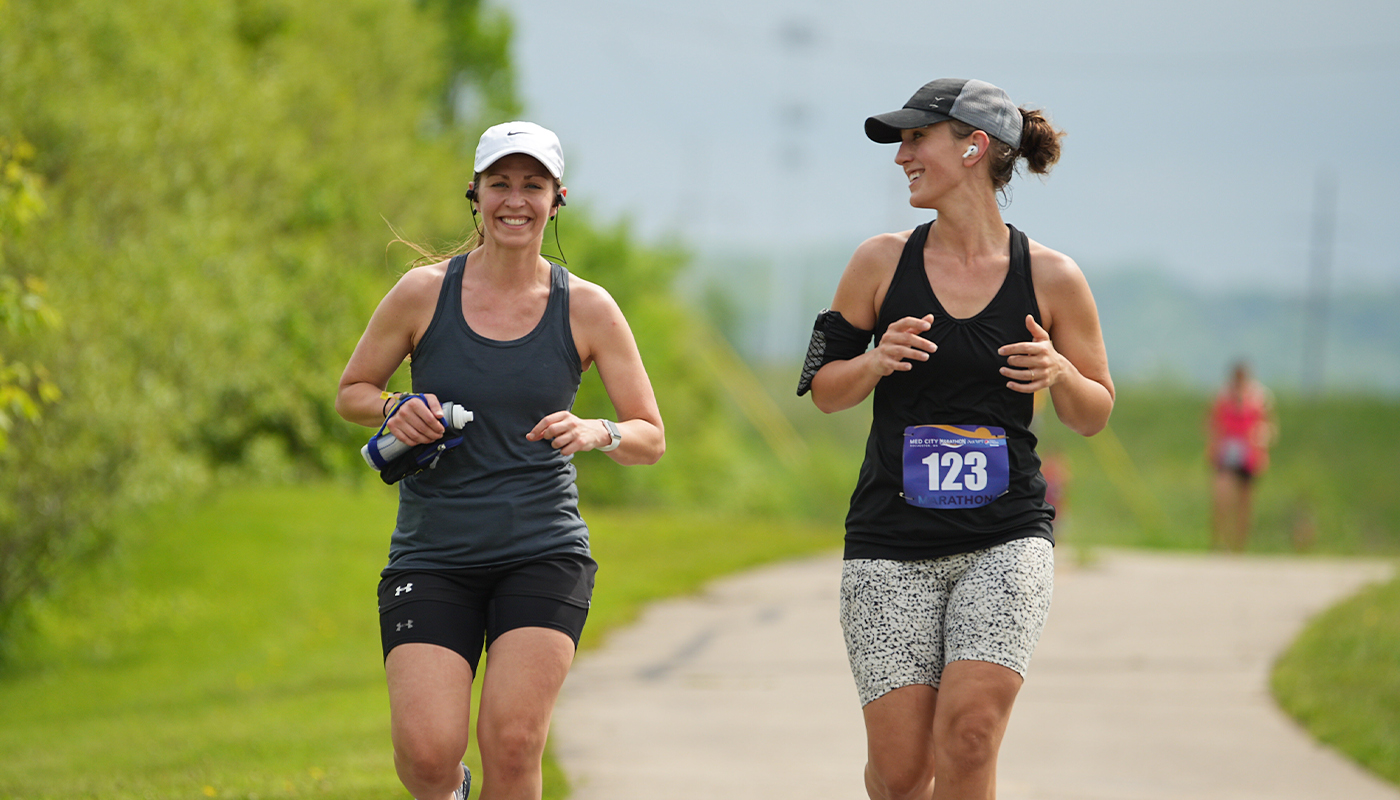 Two marathon runners smile as they run next to each other. A concrete path is behind them.