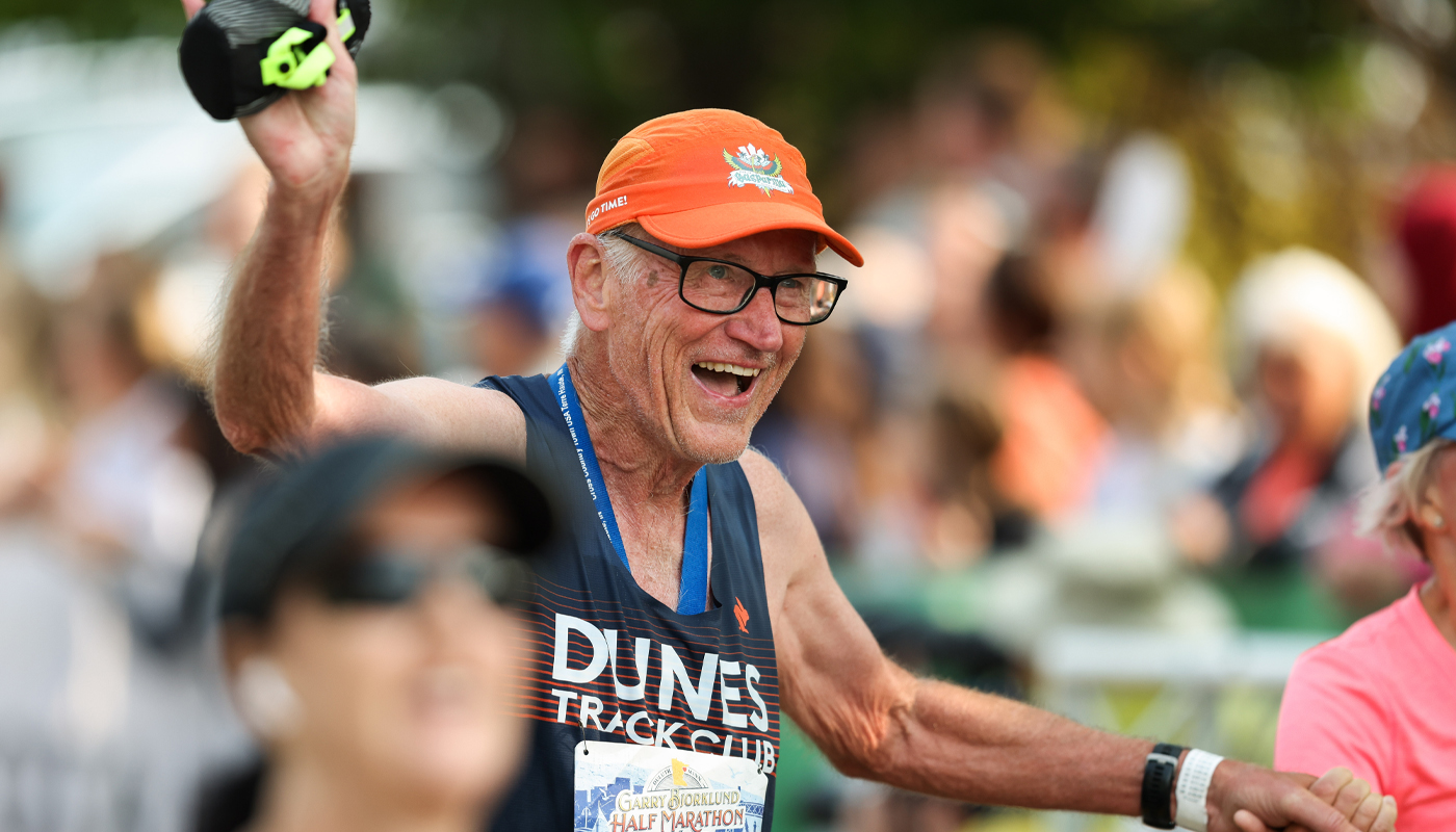 An older marathon runner holds his partner’s hand and smiles as he runs. 