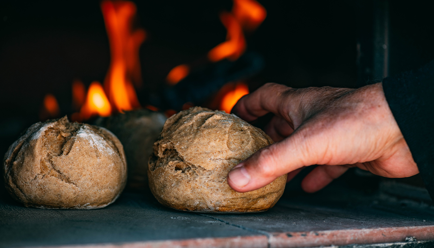 Closeup of a hand pulling bread rolls out of a brick oven with flames in the background