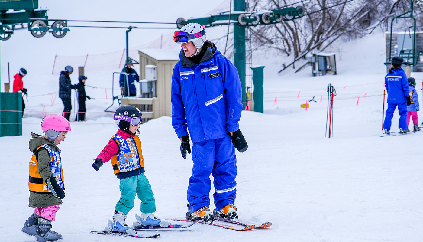 Ski instructor teaching two children how to ski with various people and a ski lift in the background