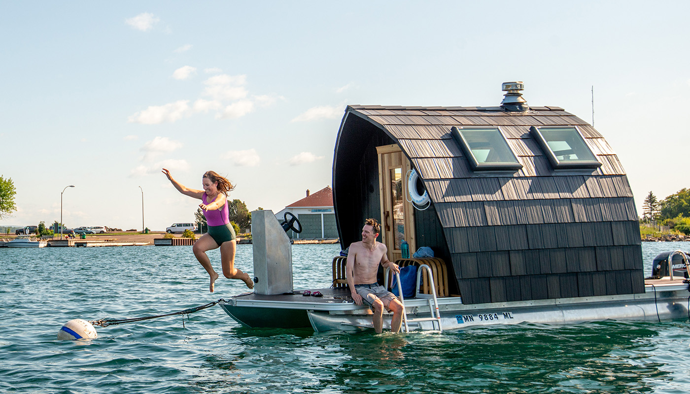 Woman jumps off of floating sauna into lake. The floating sauna has brown shingles and skylights