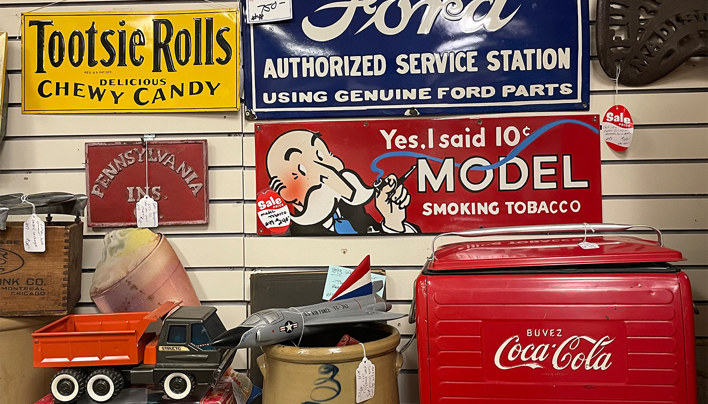 Vintage signs hang on a shop wall, advertising cars, candy and cigarettes.