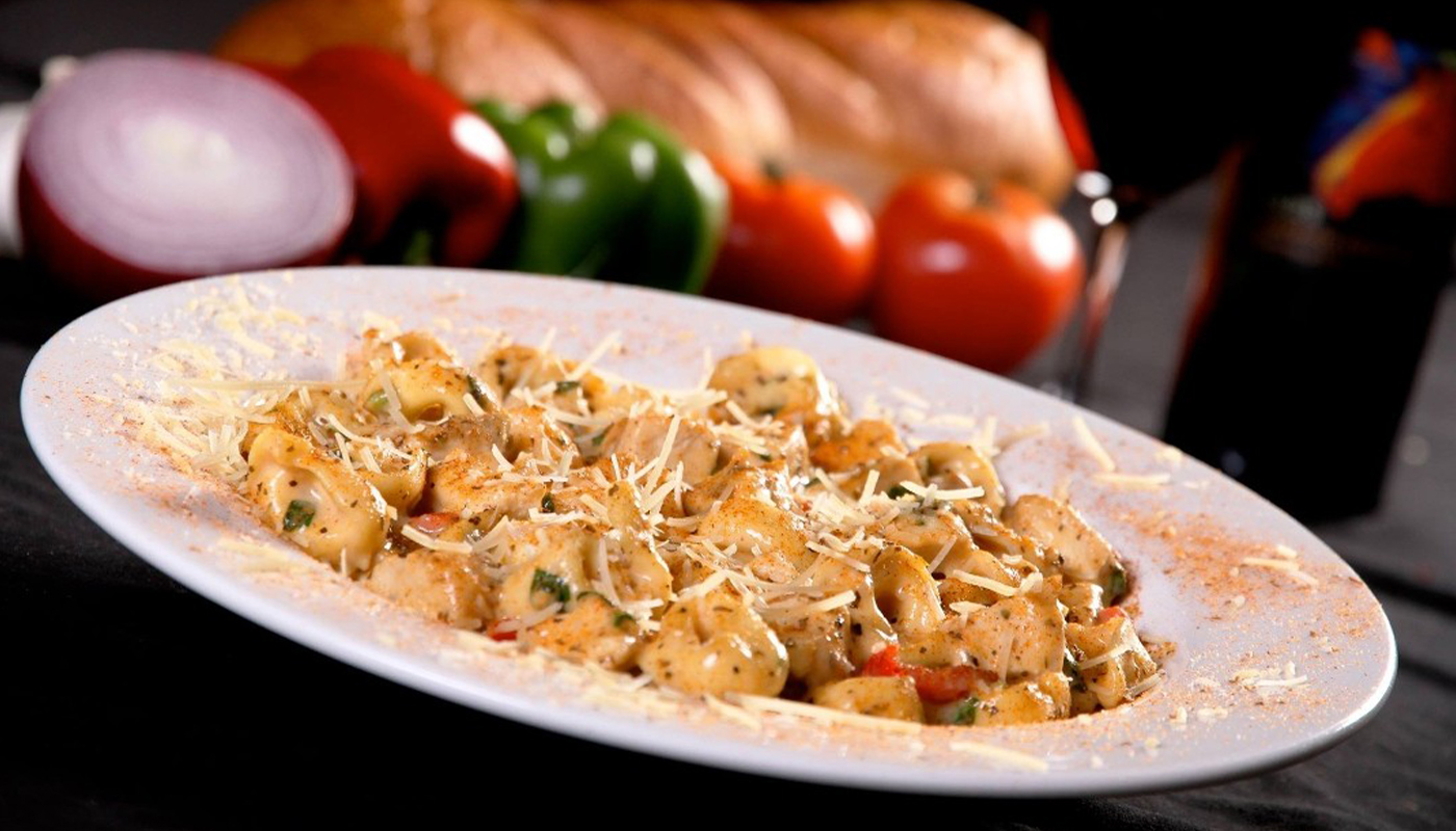 A plate of tortellini dusted with parmesan. Whole peppers and tomatoes are in the background.