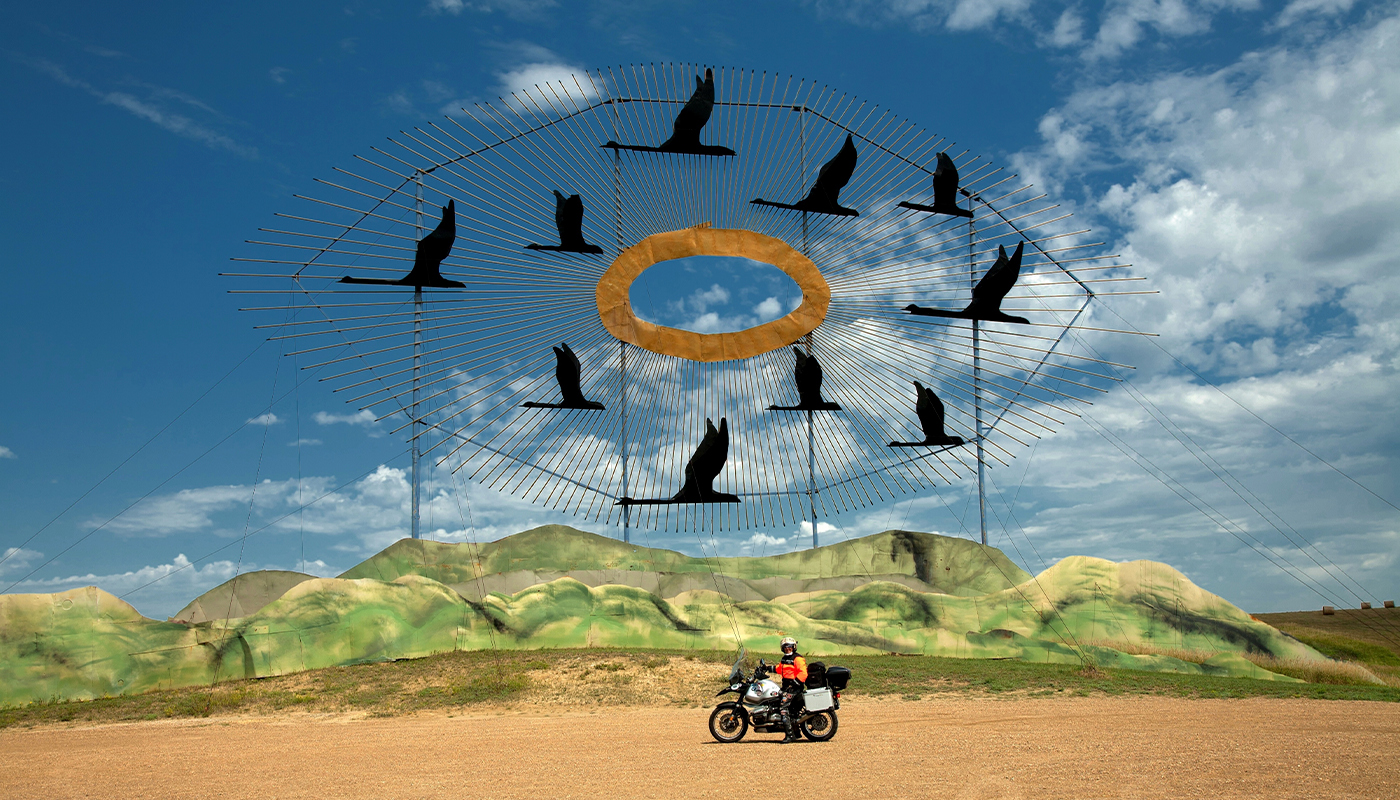 A motorcyclist poses in front of a large circular metal sculpture of birds flying across the sky.