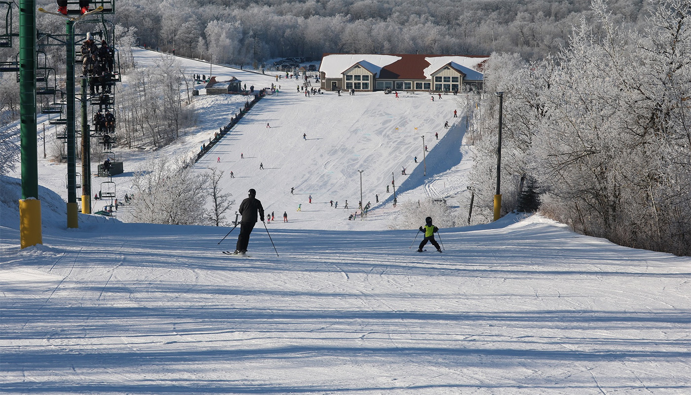 Adult and child skier on a ski slope with other various skiers and a lodge in the background