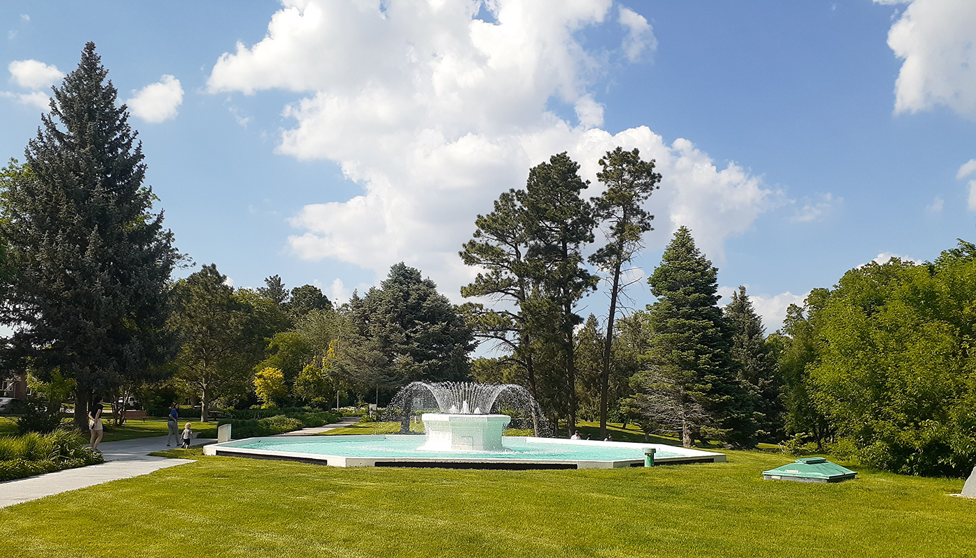 A large fountain in the middle of a manicured grassy area surrounded by green trees.
