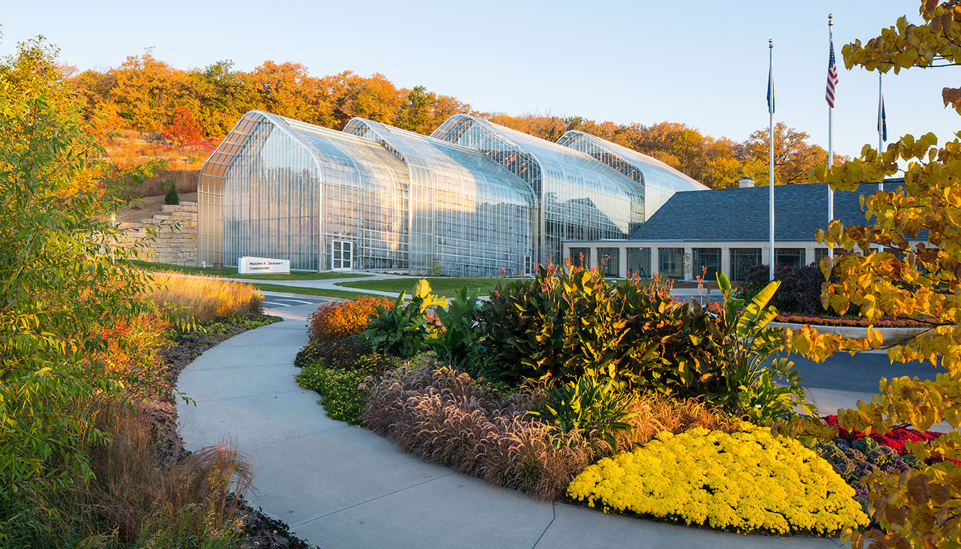 Trees and shrubs with fall foliage surround a large greenhouse. 