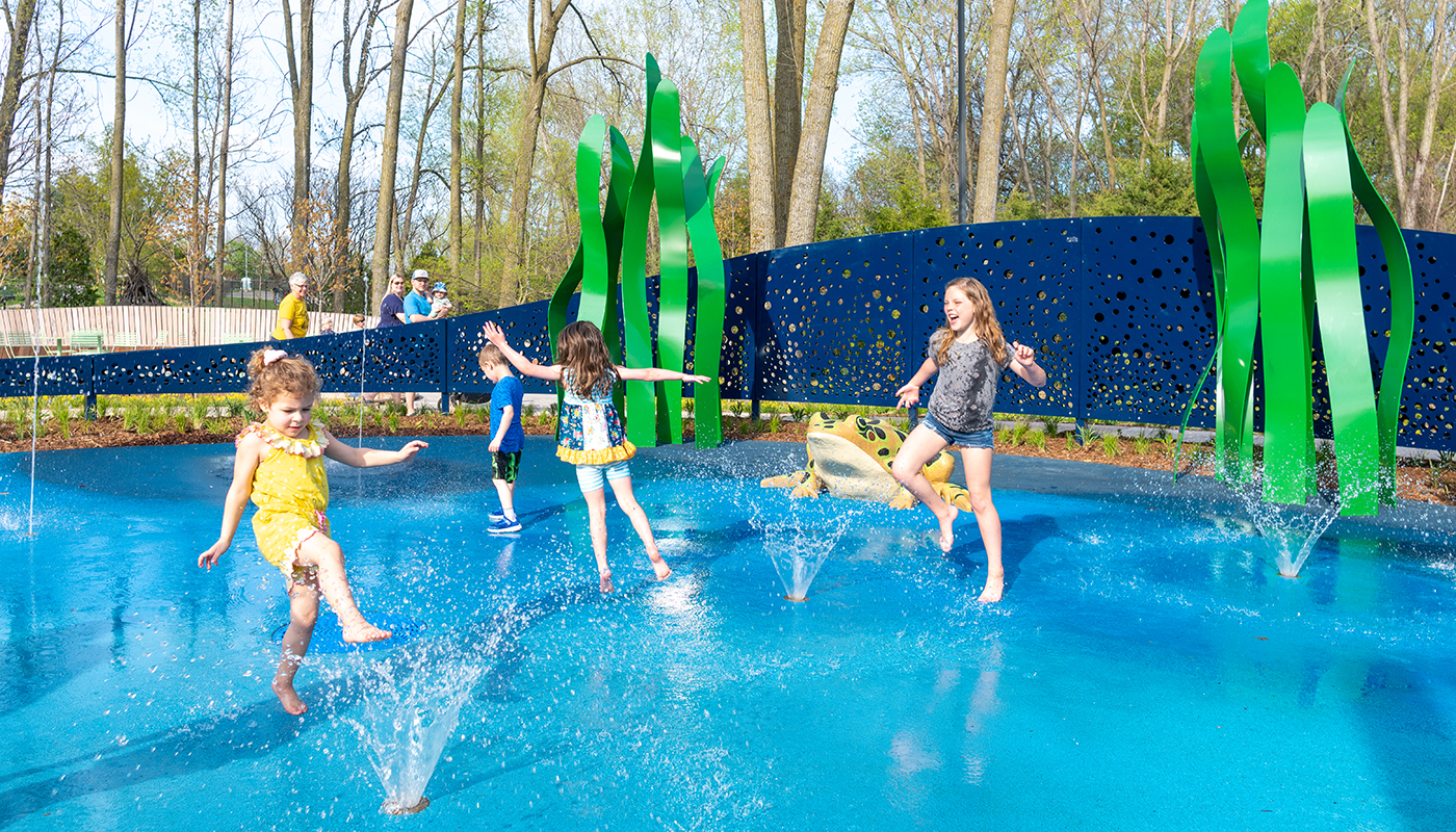 A group of children play in a splash pad in a play area. Green sculptures are in the background.