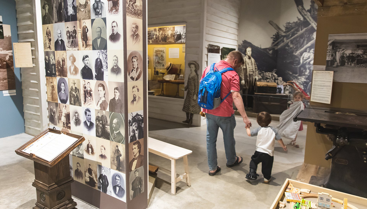 A father and child hold hands as they walk through a museum exhibit. A wall on the left is covered in portraits of people.