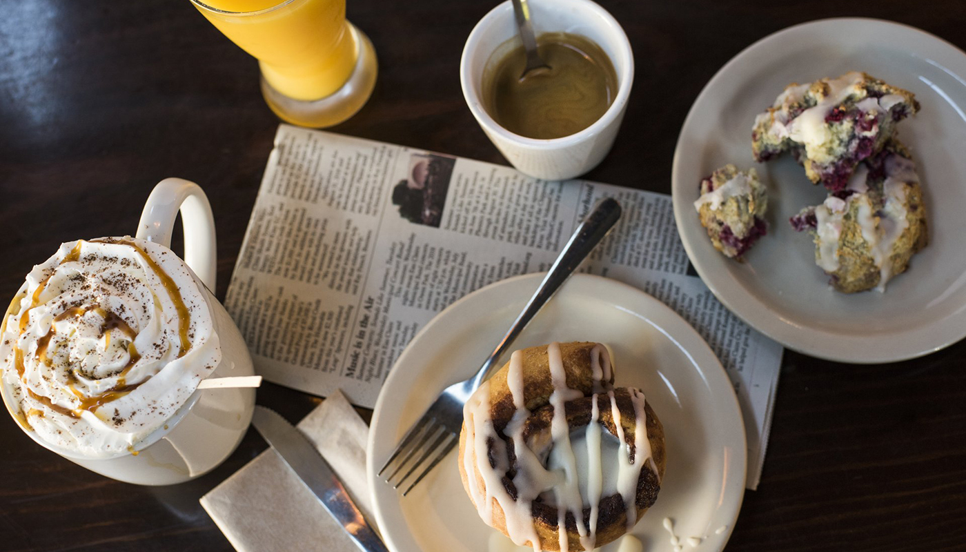 An overhead view of a breakfast including a cinnamon roll, scone, tea, orange juice and whipped cream-topped drink.