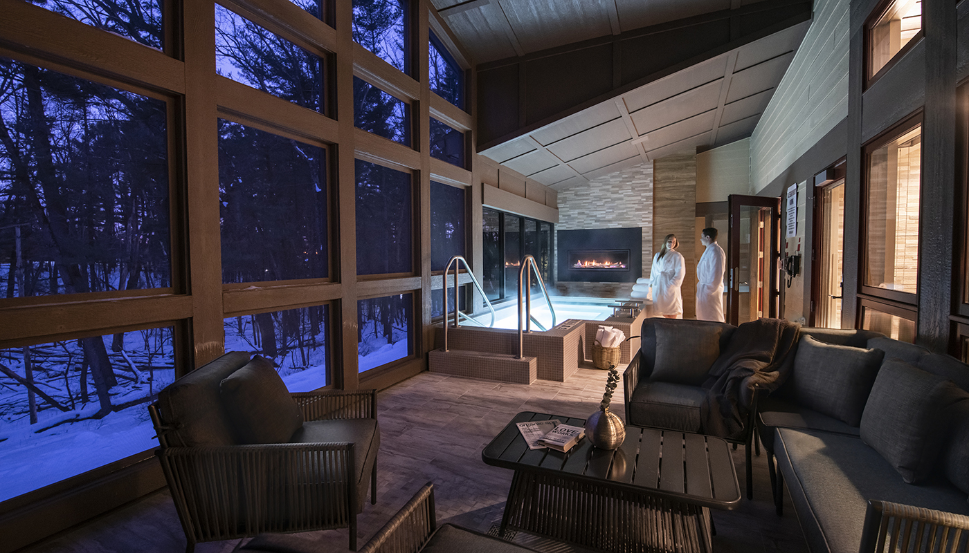 Two guests get ready to use an indoor jacuzzi in a dimly lit room. Big windows reveal a snowy scene outside.
