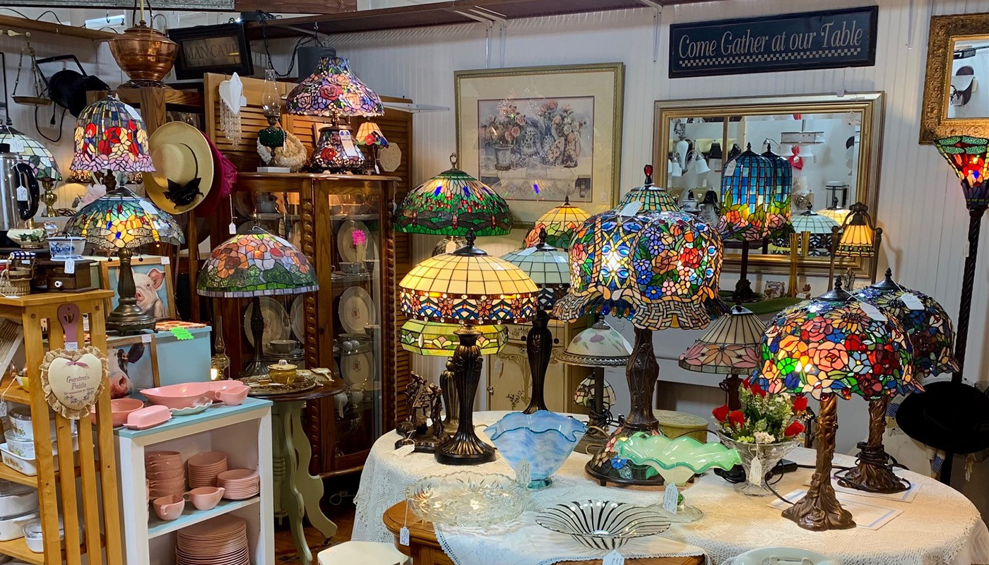 A corner of a shop is filled with antique lamps. Many of the lamp shades are made of colorful stained glass, depicting flowers and abstract designs.