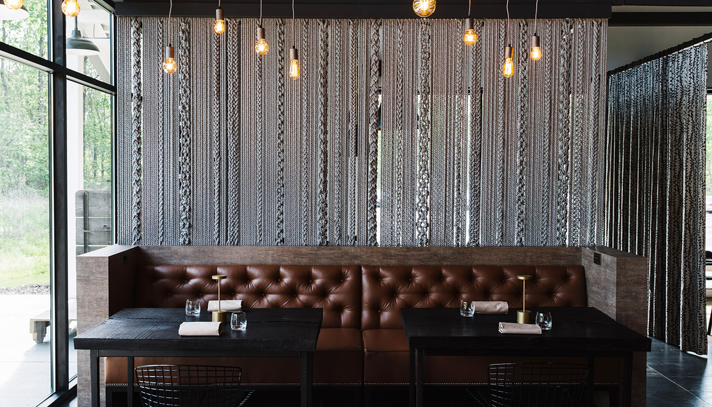 A large tufted brown leather couch in front of a wall of draped ropes. Two tables are in the foreground.