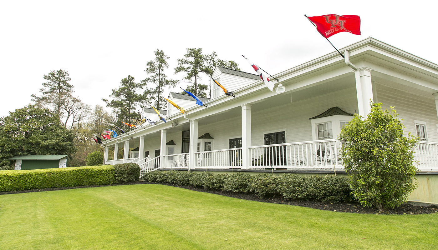 A home with white exterior and wraparound porch surrounded by grass and manicured shrubs. Multiple flags are mounted to the roofline of the house.