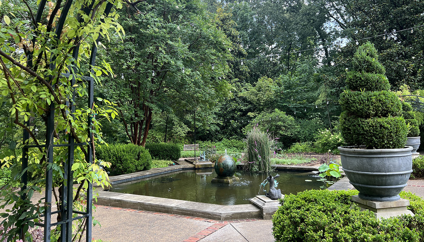 A small reflection pond with a fountain in the middle. Topiaries in containers are on the right.
