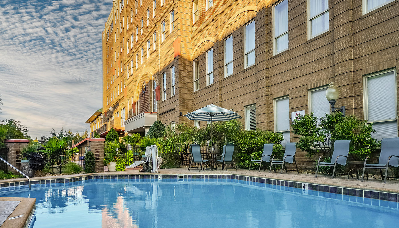 An outdoor pool in the shade of a hotel. Chairs surround the pool.