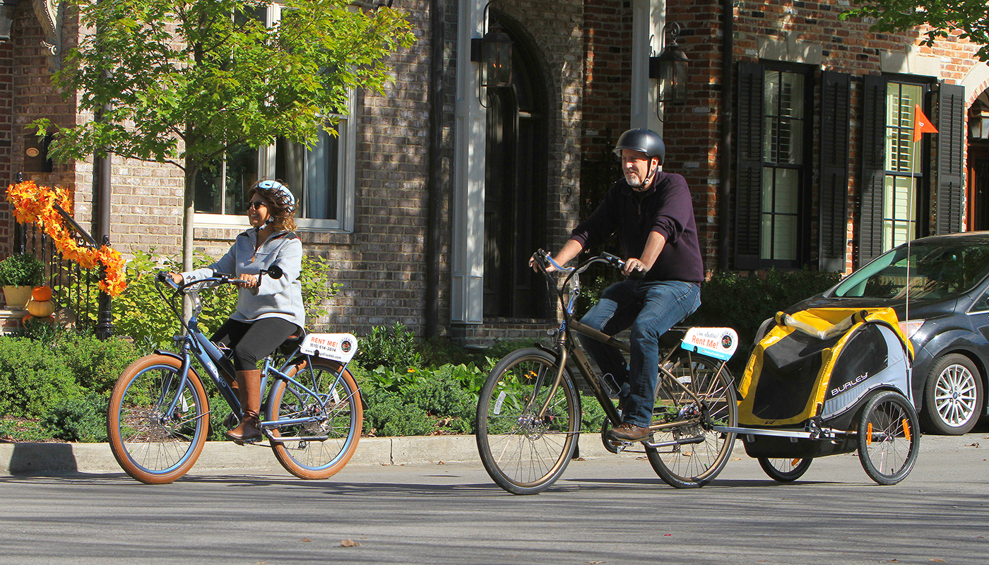 Two adults ride bikes down a neighborhood street.