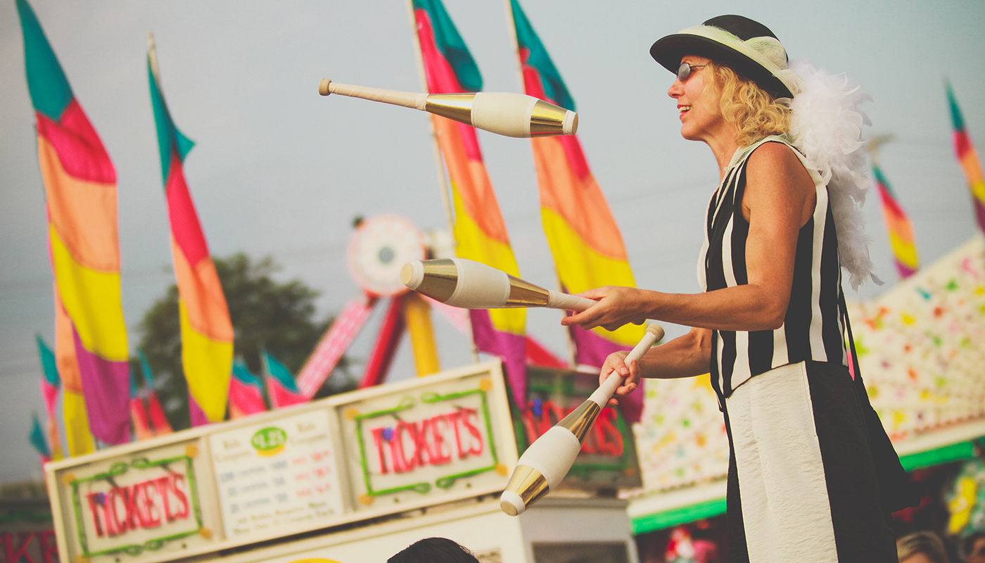 A performer juggles three clubs at a colorful fair ground.