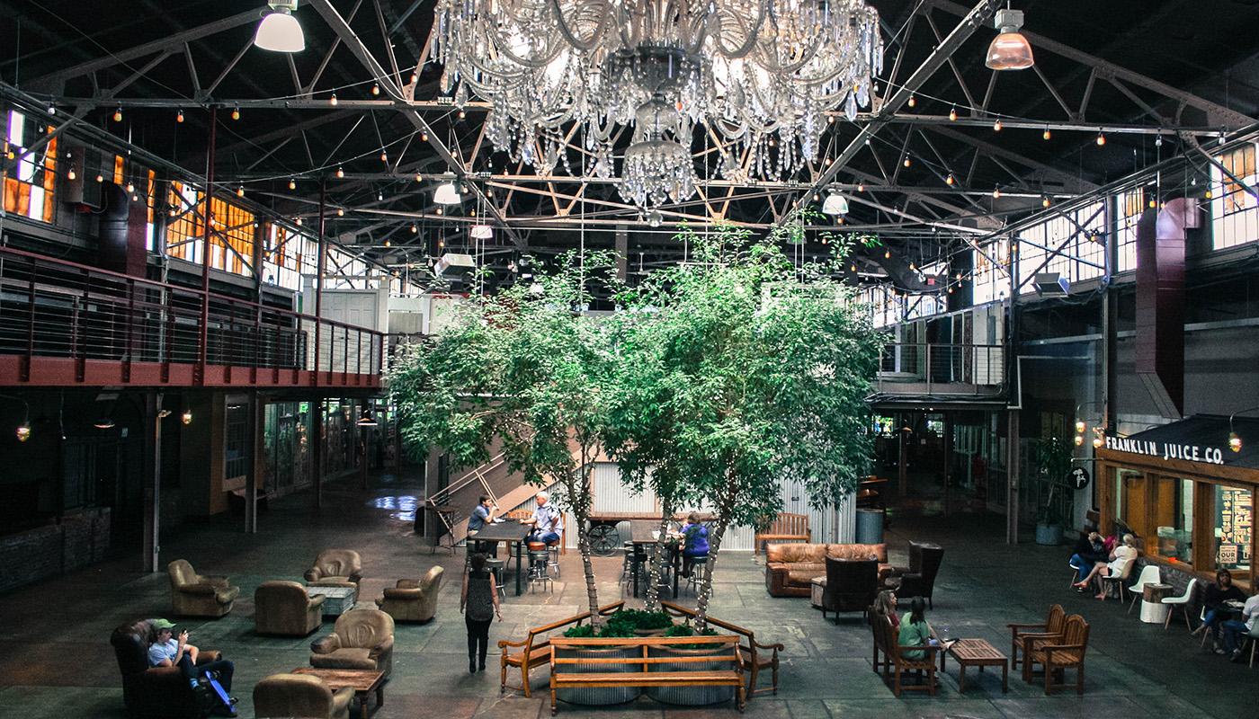 An ornate chandelier hangs above three trees inside an industrial-looking space with multiple sitting areas. 