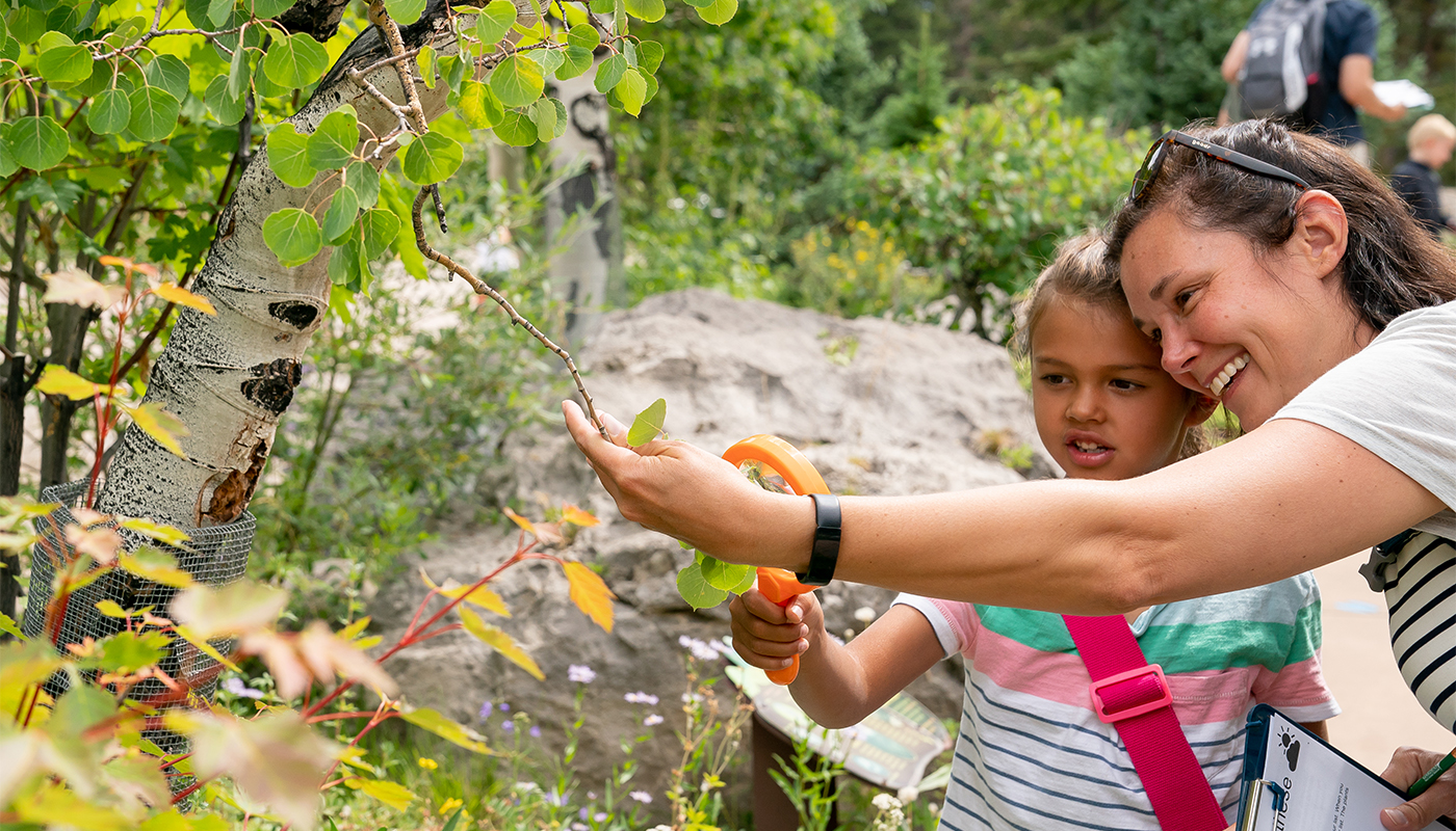 A woman holds a branch for a young girl to examine with her magnifying glass. 