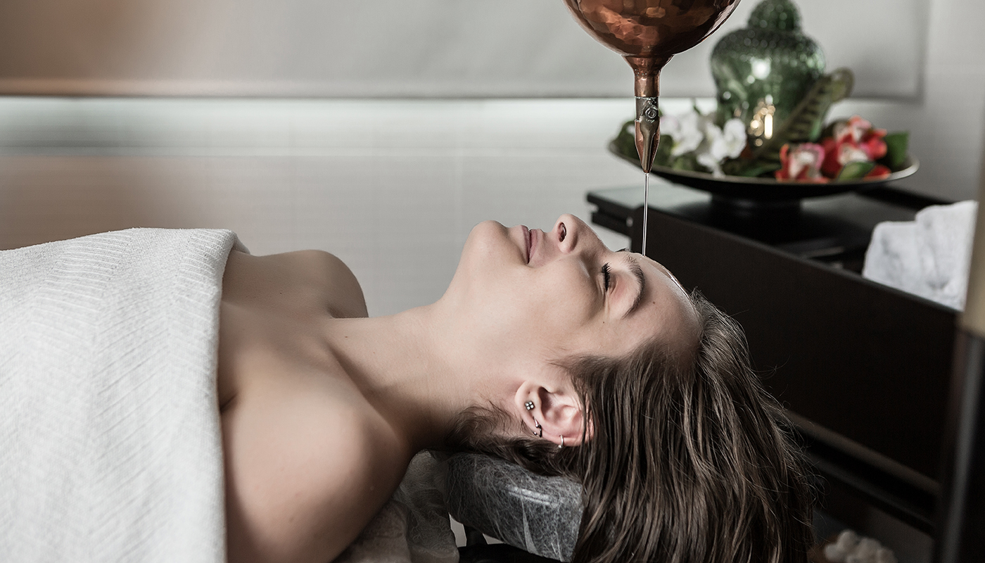 Woman lying on table and covered with a towel while warm oil is slowly poured onto her forehead