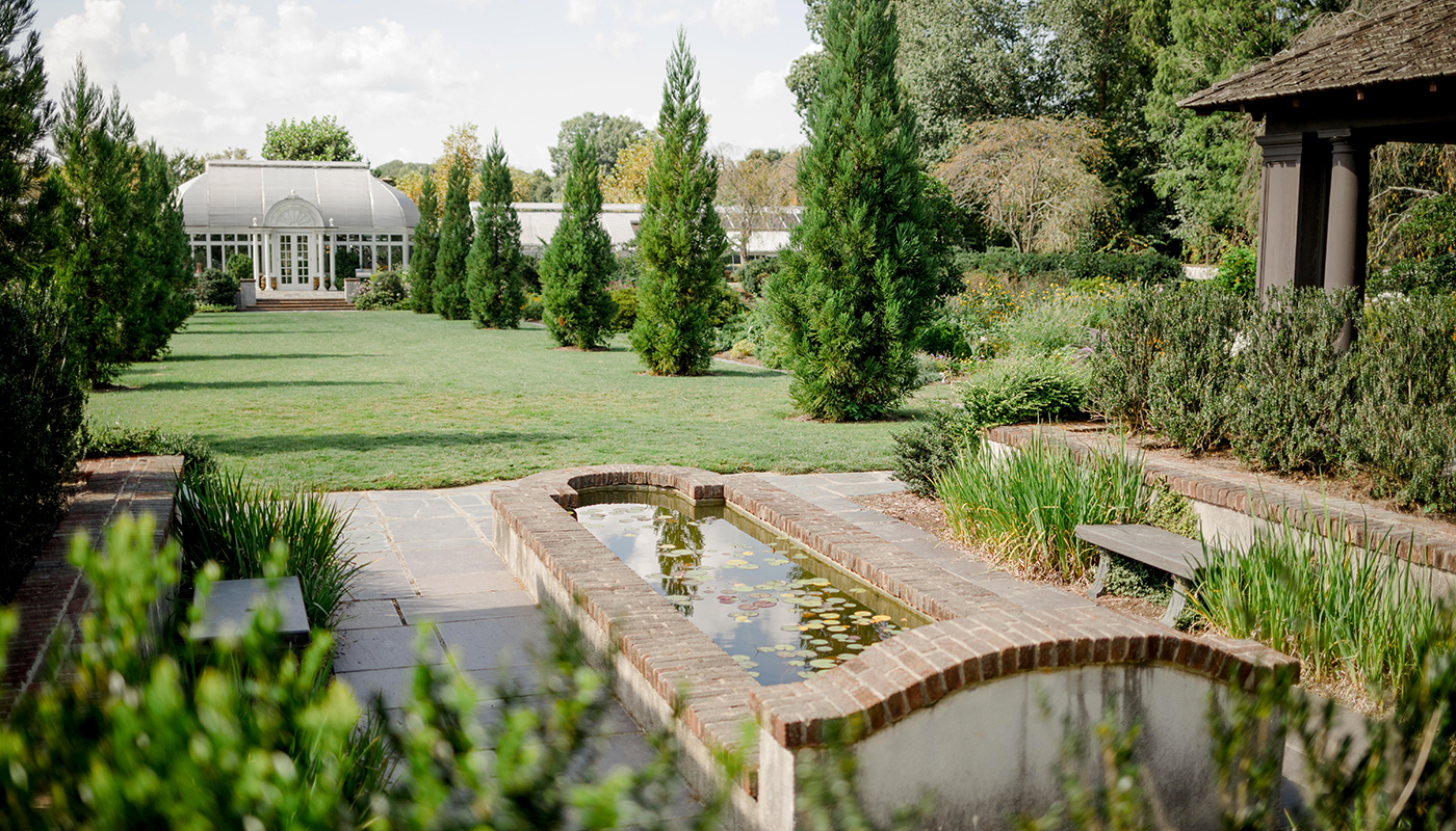 A small reflection pond with a clearing of grass behind it. A large formal greenhouse is in the background.