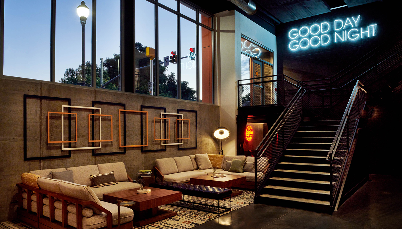 A hotel lobby with multiple sectional sofas and tables. A neon sign hangs above a staircase that reads “GOOD DAY GOOD NIGHT.” Large windows are on the left.
