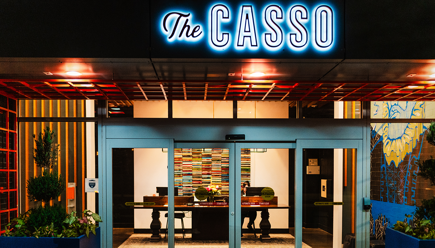 Exterior view of a softly  lit restaurant with a neon sign that reads “The CASSO” in white, black and blue.