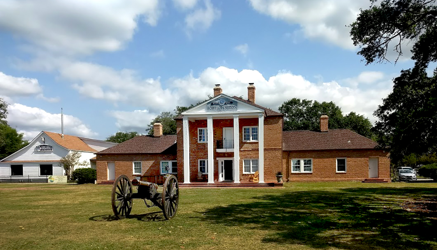A cannon rests in front of the Fort Johnston Visitor Center, a large brick building.
