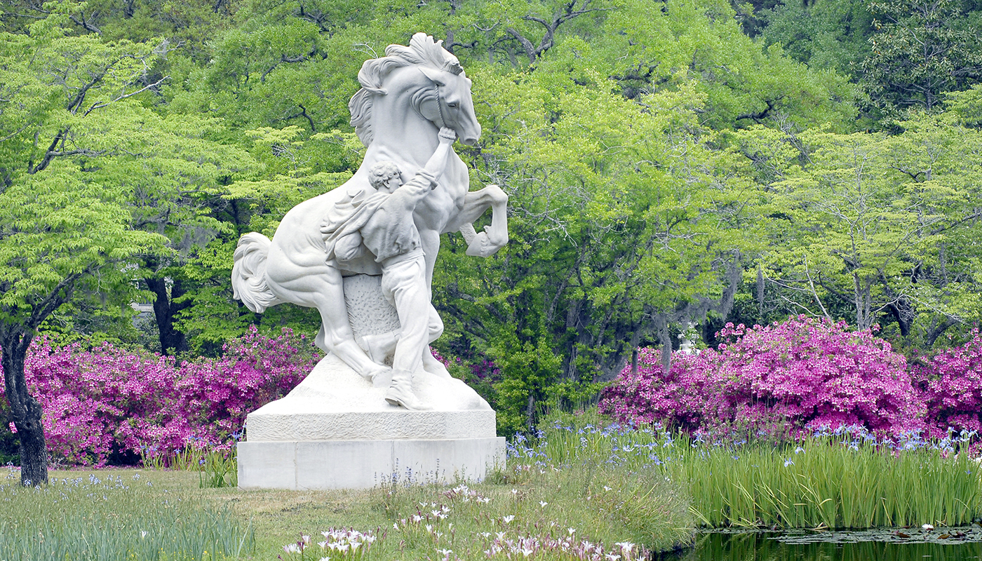 A statue of a man holding a rearing horse by its bridle beside a pond. Pink azaleas and trees are in the background.
