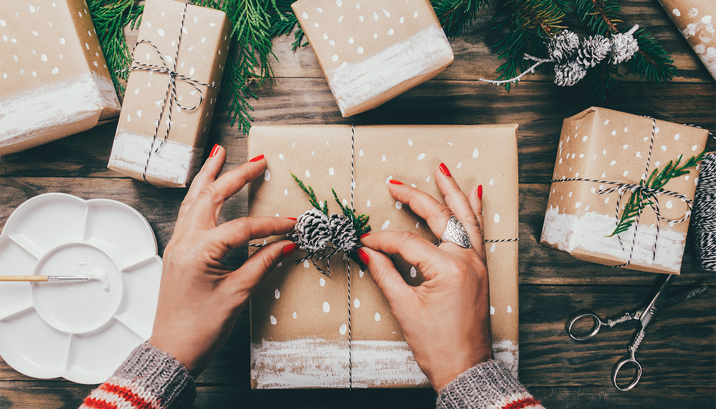 Woman´s hands wrapping Christmas presents on brown paper decorated with painted snow, fir branches and pinecones on a rustic wooden board. Top view