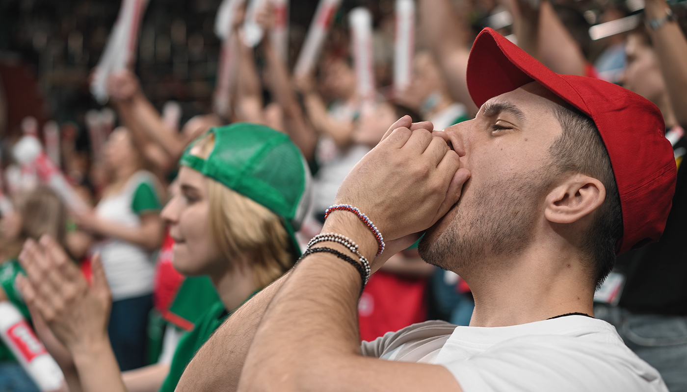 A man in a red cap shouts through his cupped hands in a raucous crowd at a sports game.