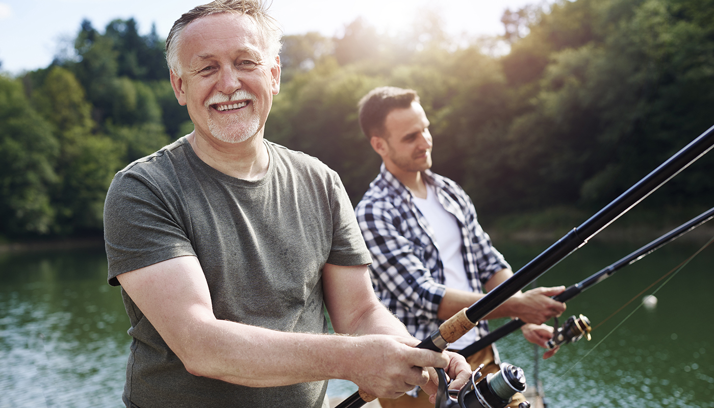 Two men are fishing on the dock, one older and one younger. The older man smiles at the camera.