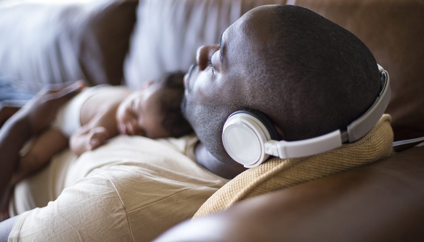 A man listens to his headphones as his infant child sleeps on his chest.