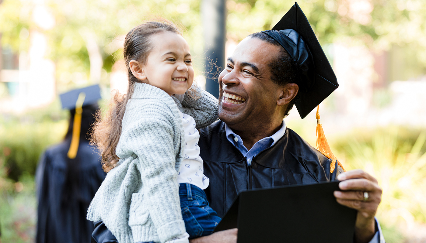 A dad in graduation regalia holds and smiles at his young daughter. 