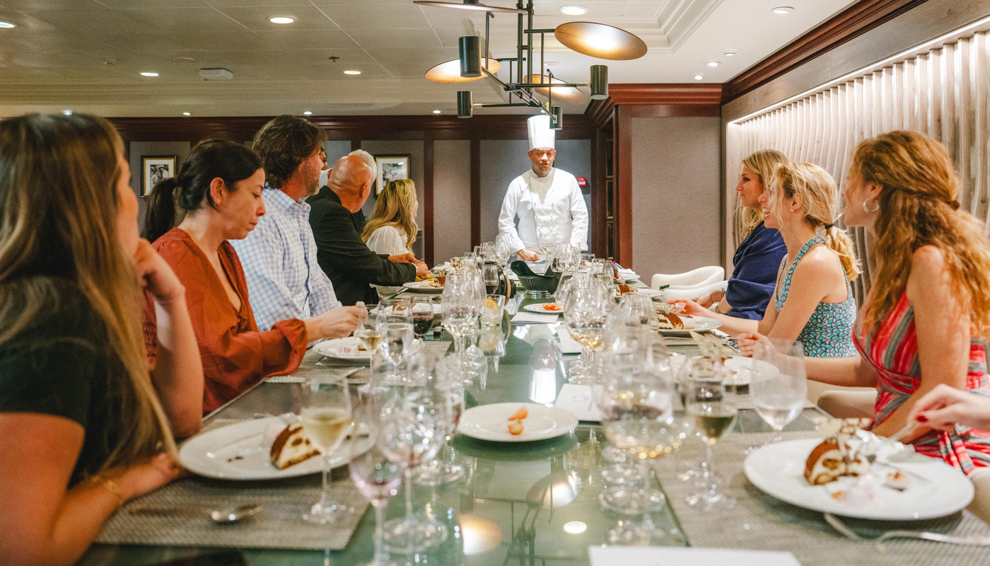 Group of people at table with chef at the head about to eat on board cruise ship.