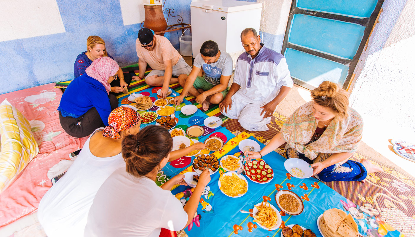 A group of tourists sitting on the ground eating with locals at a makeshift table.