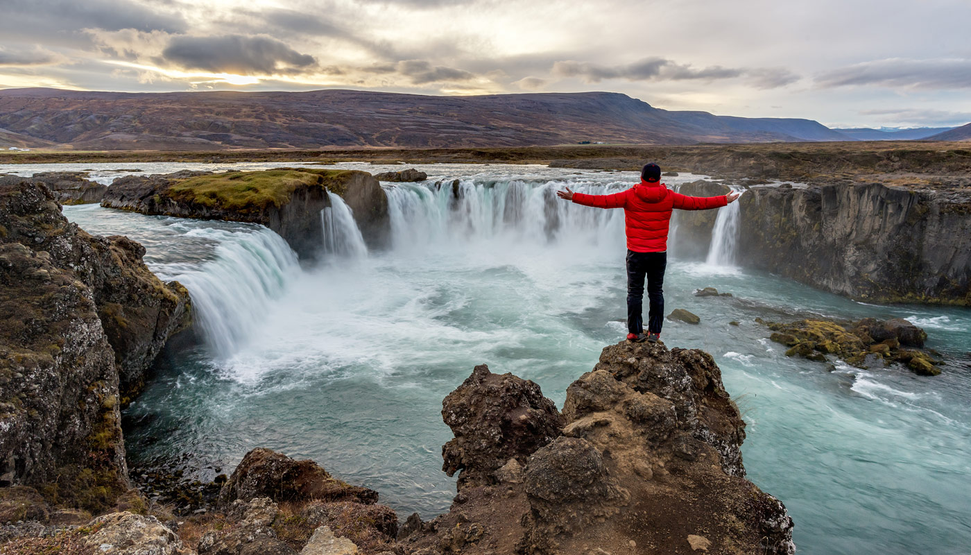 Picture of a man in a red puffy jacket overlooking a waterfall in Iceland from on top of a cliff.