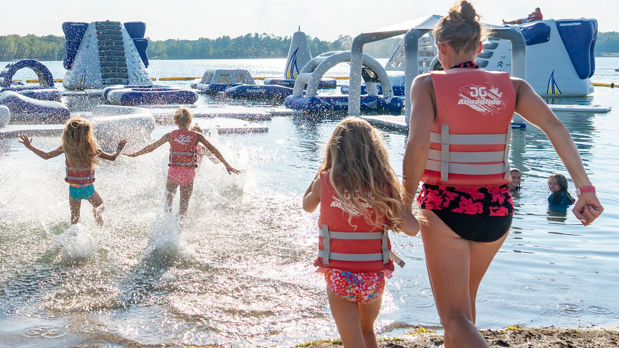 A woman and 3 kids wearing swim suits going into a lake towards an inflatable course.
