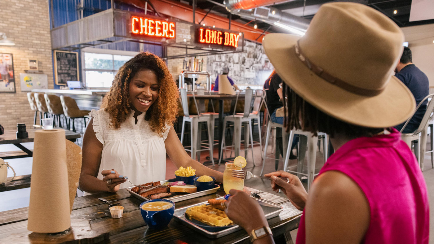 Two women at a bar/restaurant with trays of food in front of them on the table with a bar with neon lights saying Cheers and Long Day ablove the bar.